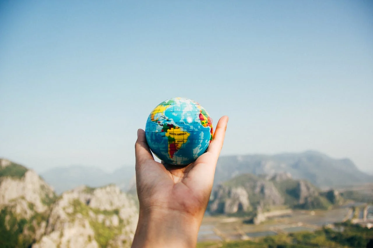 hero-img-02 Hand holding a globe against a mountain background symbolizing travel and exploration.